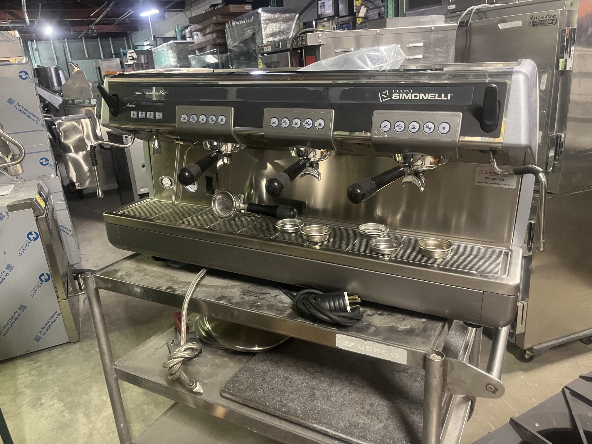 Nuova Simonelli Aurelia V 3-group espresso machine front view showing brew groups, steam wands, and portafilter baskets on a stainless steel cart