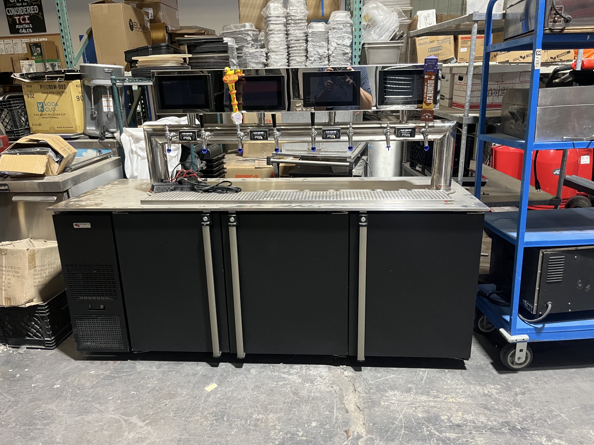 PourMyBev self-pour beverage station with 8 chrome taps, 4 touchscreen stations, and three-door Micro Matic refrigerated base — front view in warehouse