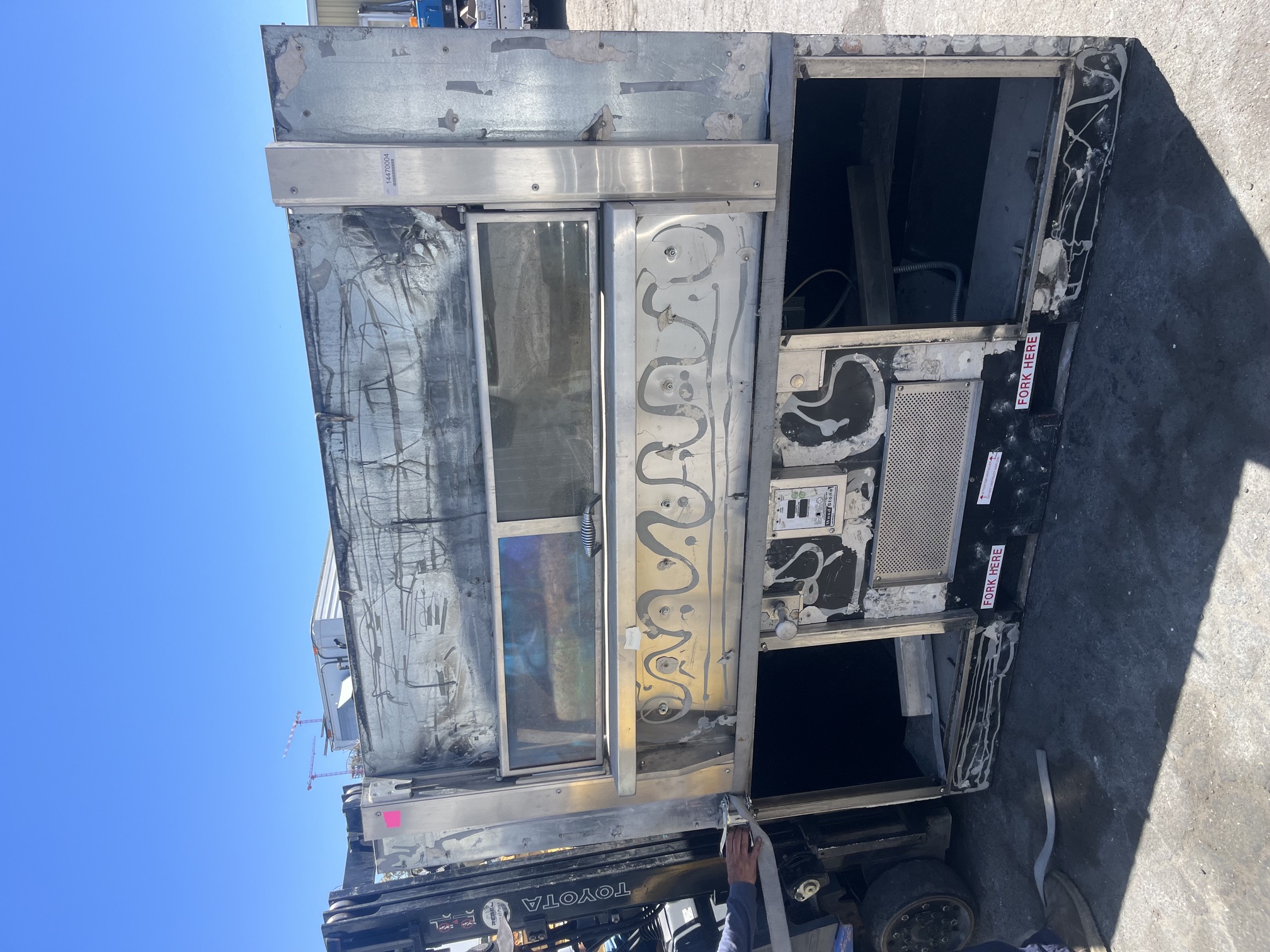 Front view of a used Wood Stone FD-9690 stone hearth gas-fired pizza oven on a forklift, showing heavy cosmetic wear, dual viewing windows, and Fork Here labels on the base